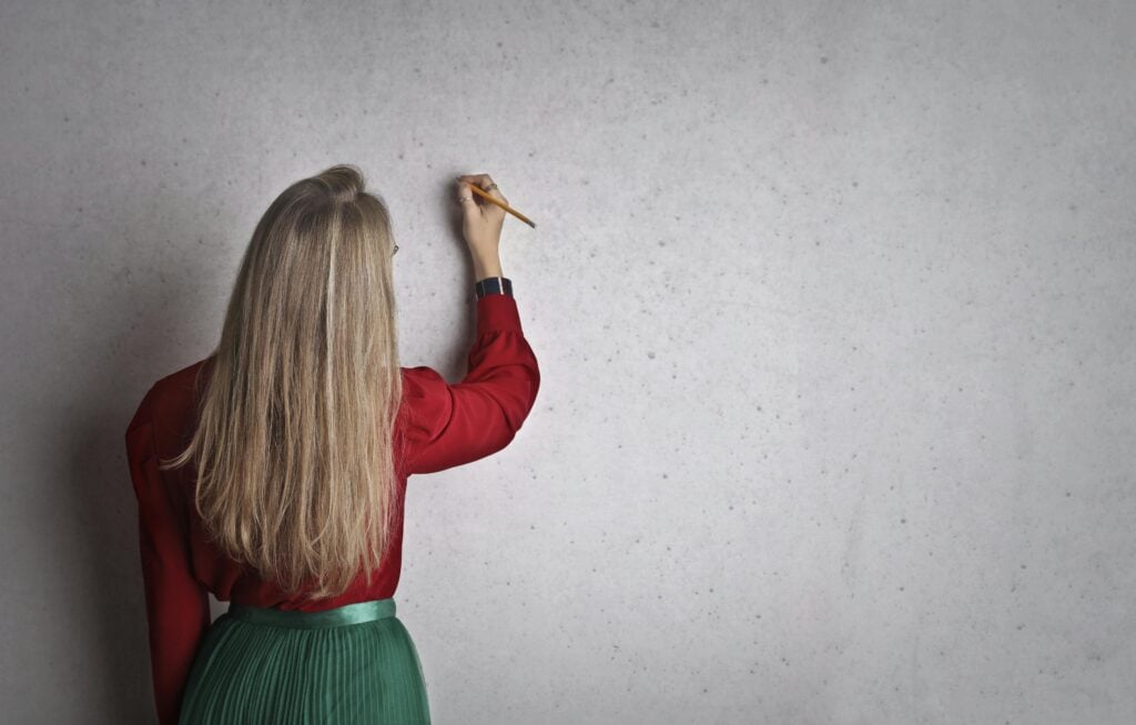 A woman in a red blouse writing on a concrete wall, viewed from behind.